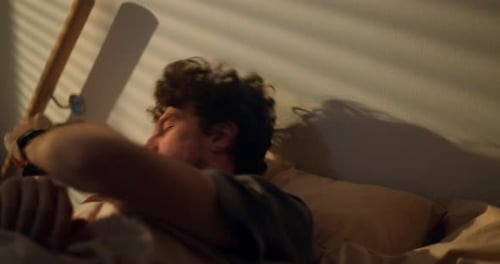Ein brünetter Mann mit lockigem Haar stellt seinen Wecker für eine späte Zeit ein und ruht sich weiterhin in seinem Bett aus