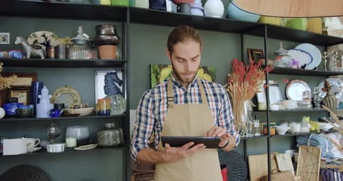 Smiling man seller in apron using tablet device and then looking at camera while working in own shop