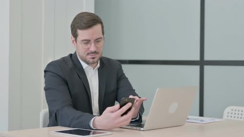 Man Works on Laptop and Phone in Office