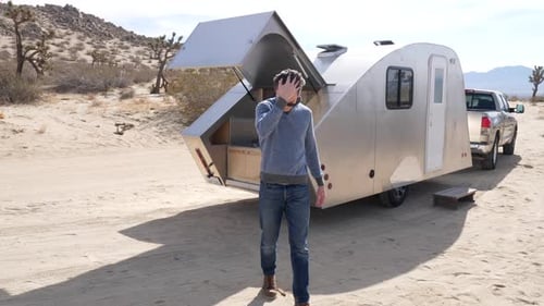 Slow motion shot of a handsome white man on a desert camping trip next to a metal travel trailer tin