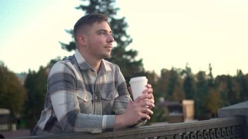 A Young Man with a Glass of Coffee in His Hand A Man Near the Fence on the Embankment with a Hot