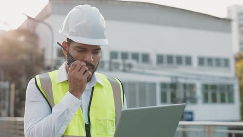 Porträt eines männlichen Ingenieurs mit Laptop, der über ein tragbares Radio gegen ein Gebäude spricht