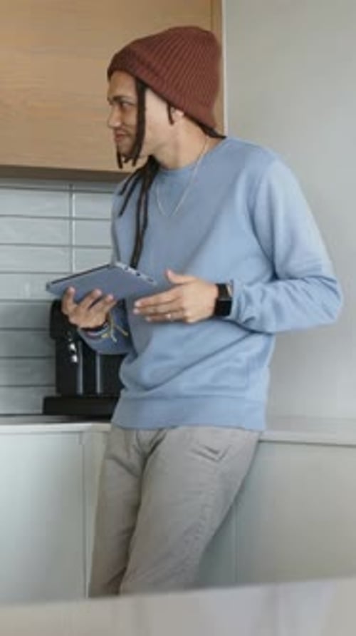 Vertical video: Glancing at mug, man in beanie with tablet in kitchen, reaching and sipping coffee