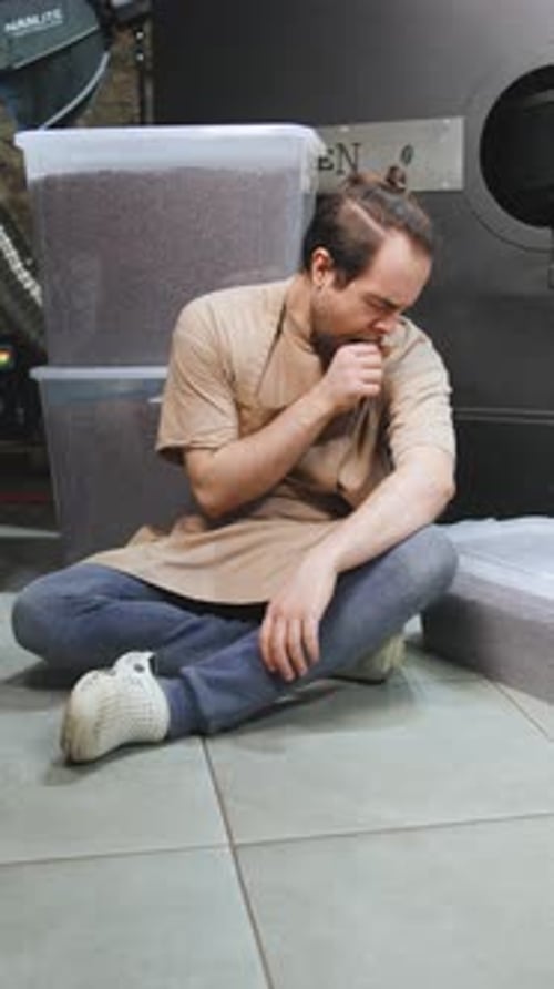 Sleepy Barista Sits on the Floor Yawns Near Coffee Bean Containers in a Roasting Area During a Busy
