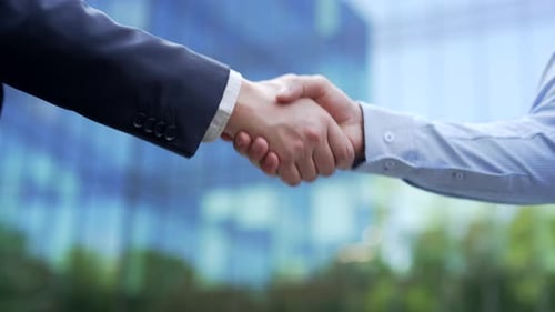 Close up of two male professionals shaking hands outdoors with a blurred office building