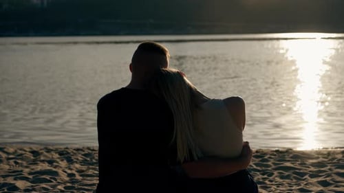 Silhouette of couple in love sitting on the sandy bank of the river admiring the setting sun