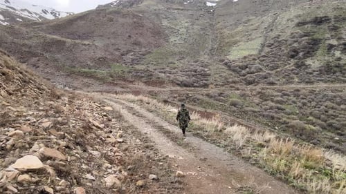 Camouflage Figure Walks Mountain Path Through Rural Landscape