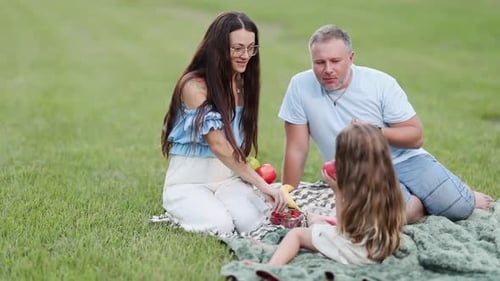 A Family Sits Together on a Picnic Blanket in a Green Park Sharing Fruit and Enjoying a Calm Summer