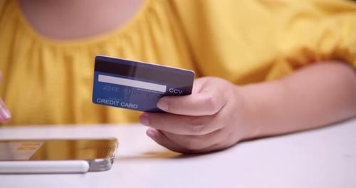 A woman dressed in yellow holds a credit card while checking transactions on her cellphone. Online p