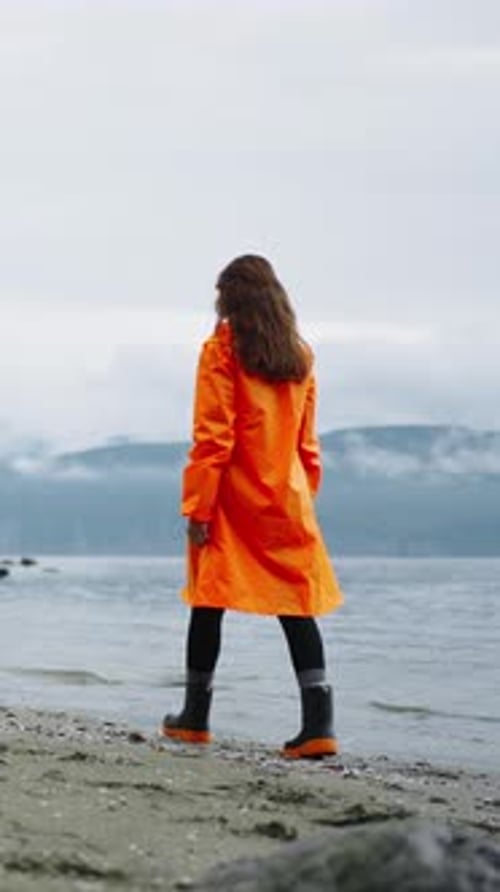 Woman Walks on Beach in Orange Raincoat and Boots