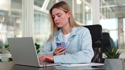 Stylish Lady Gazing at Display of Phone While Typing on Laptop in Modern Office