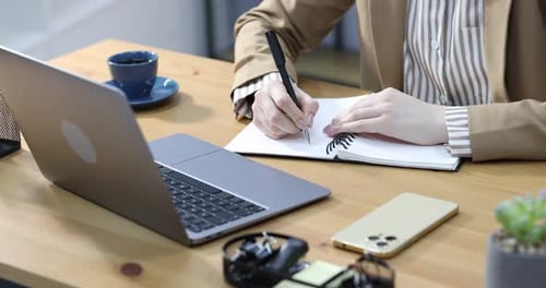 Woman writing and working with laptop at wooden table indoors, closeup
