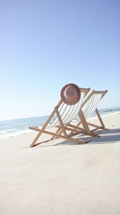 Vertical video: Beach scene with two empty lounge chairs and hat, relaxing by sea