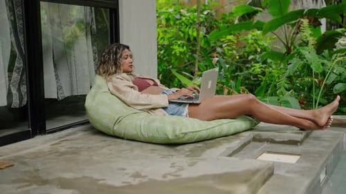 Relaxed Biracial Woman Works on Laptop Lounges on Bean Bag By Pool in Tropical Garden Freelancer