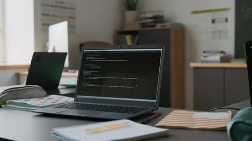 Desk with Two Laptops Displaying Code in Modern IT Classroom with Blackboard