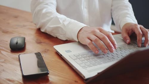 A Young Adult Man in a White Shirt Works on a Laptop with a Wireless Mouse There is a Smartphone