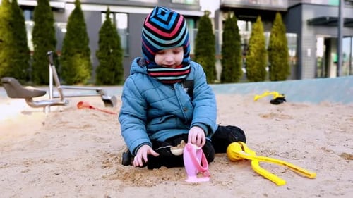 Young Child Plays in Sand Using Toys at a Playground on a Chilly Day Dressed Warmly in a Blue Coat
