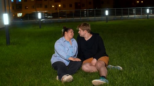 Happy romantic couple sitting on the green grass near building and street lamps