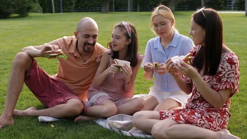 Happy Family are Sitting on the Grass in the Park and Biting Sandwiches Together