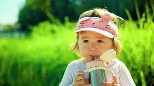 Adorable american child girl drinking from kids' water bottle in a park against lush green bushes -
