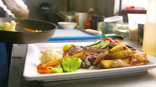 Chef using metal tongs to place grilled vegetables on plate with steak and potatoes