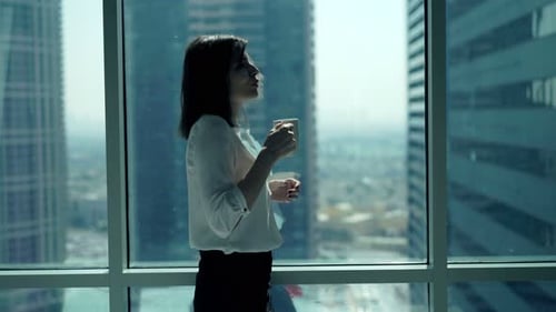 Young Businesswoman Relaxing and Drinking Coffee Standing by Window in Office 30s
