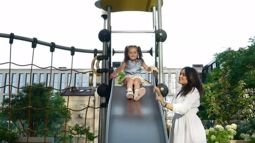 Young Girl Enjoys Sliding Down Playground Slide While Mother Supervises in City Park