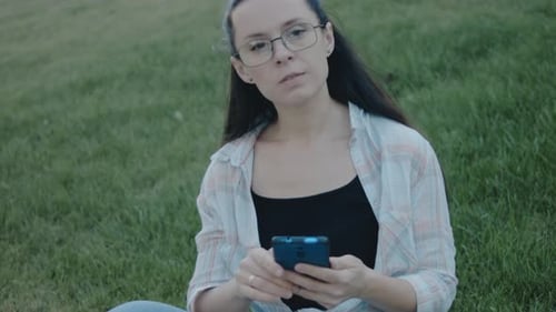 Young Woman with Eyeglasses Sitting on Green Grass Using Mobile Phone Looking at Screen Texting