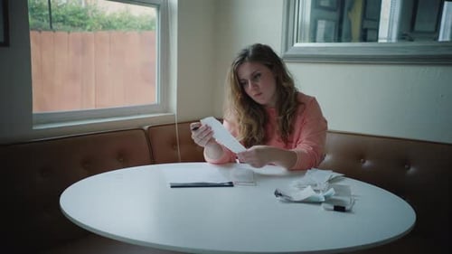 Handheld Shot of Woman Organizing Receipts on Corner Kitchen Table By Window, Wearing Pink Sweater