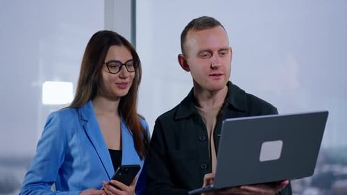 Office employees stand at the window and are focused on the laptop in man’s hands.