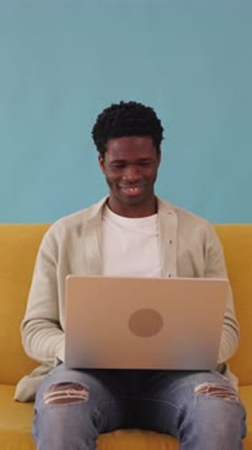 Vertical Video of Young African Man Sitting on a Yellow Couch Using Laptop