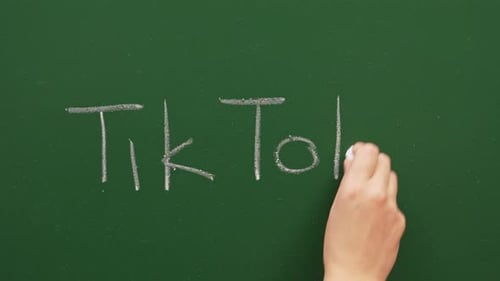 A Hand Writes Tik Tok On A Green Chalkboard