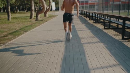 Back View of Shirtless Man Running on Pathway in Sunny Urban Park