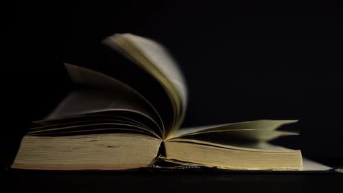 Slow motion, close up of an old, vintage, open book with pages turning on the wind, on a dark room w