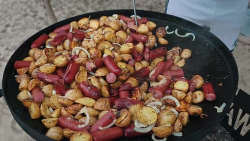 Stirring Fried Potatoes and Sausages on a Large Disc Grill