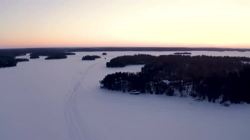 Snowmobiles traveling down a trail as camera ascends to reveal the sun setting on the horizon of a f