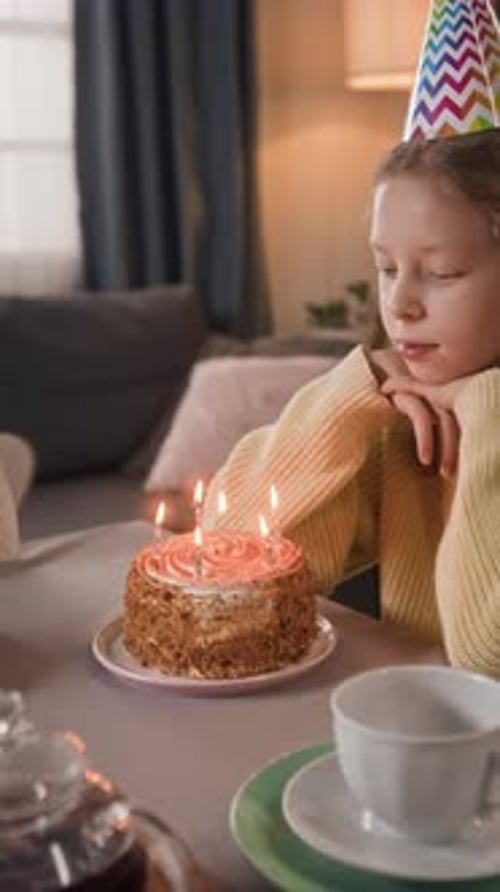 Vertical of Gen Alpha Girl with Party Hat Blowing out Candles on Festive Cake