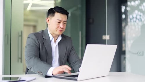 Frustrated asian man having problems with software or service on laptop while sitting at workplace