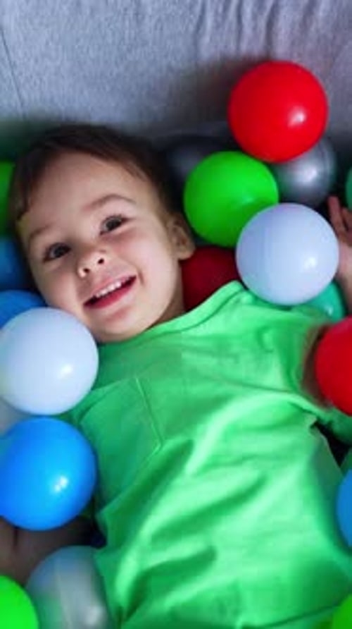 Happy smiling toddler lies in the dry pool. Charming Caucasian baby boy in a pile of colorful balls.