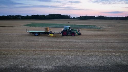 Tractor collects square bales of straw with bale picker from a recently harvested field and places t