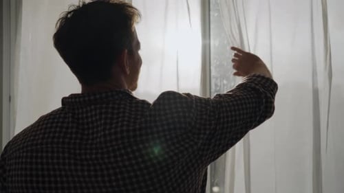 Young man opening white window curtain looking outside to city in early morning sunlight or during