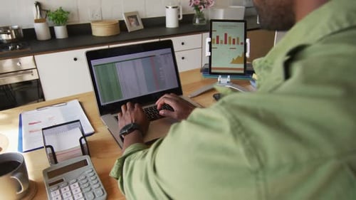 African american man sitting at table in kitchen, using laptop and tablet