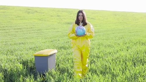 A woman in a yellow protective suit stands in the middle of a green field and holds a globe