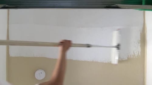 Mãos de um homem pintando o teto de branco com rolo de pintura dentro da casa