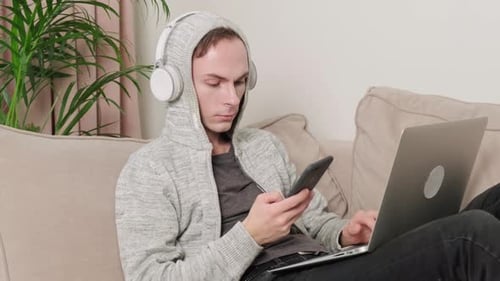 A Man with a Mobile Phone in His Hands Works with Headphones Lying on the Couch