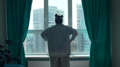 Rear view of a woman with folded hands looking out the window in domestic room.
