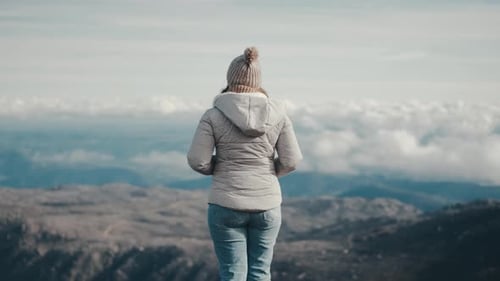 Young thoughtful woman in a gray jacket, hat, and scarf contemplates a stunning mountain landscape o
