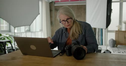 Mature Adult Female Photographer sitting in her Studio checking Camera and using Laptop