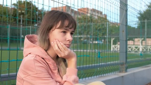 Pensive Girl Sits by Chain-Link Fence
