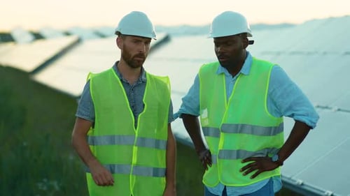 Junge afroamerikanische und kaukasische Ingenieure stehen in besonderer Uniform auf dem Solarpark und diskutieren